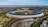 An aerial view of a large, circular building surrounded by greenery. The structure is sleek and modern, with a minimalist design that includes extensive use of glass and solar panels on the roof. The building is situated in a landscaped environment with trees, pathways, and a parking area. The backdrop includes a sprawling urban area and mountains in the distance under a clear blue sky.