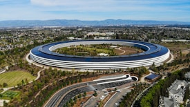 An aerial view of a large, circular building surrounded by greenery. The structure is sleek and modern, with a minimalist design that includes extensive use of glass and solar panels on the roof. The building is situated in a landscaped environment with trees, pathways, and a parking area. The backdrop includes a sprawling urban area and mountains in the distance under a clear blue sky.