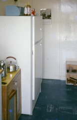 A small kitchen space contains a white refrigerator, a wooden cabinet with a metal kettle and toaster on top, and a calendar hanging on the wall. The floor is blue, and several items are placed on a shelf next to the refrigerator.