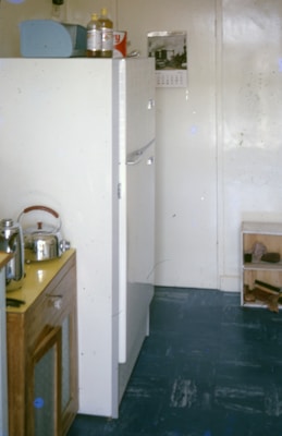 A small kitchen space contains a white refrigerator, a wooden cabinet with a metal kettle and toaster on top, and a calendar hanging on the wall. The floor is blue, and several items are placed on a shelf next to the refrigerator.