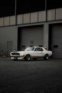 A classic white Mustang car is parked in front of a large industrial garage with horizontal sliding doors, marked by the number 12. The setting appears to be a quiet, empty parking lot with muted lighting, highlighting the sleek lines and vintage style of the vehicle.