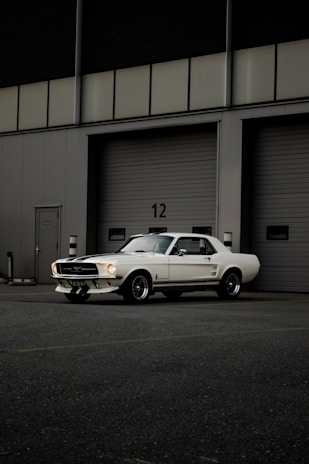 A classic white Mustang car is parked in front of a large industrial garage with horizontal sliding doors, marked by the number 12. The setting appears to be a quiet, empty parking lot with muted lighting, highlighting the sleek lines and vintage style of the vehicle.