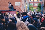 A group of people are gathered in an outdoor alleyway, raising their fists in a sign of protest. Many are wearing masks, and some wear bandanas or hats. The background shows a brick building with a sign and some greenery.