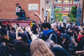 A group of people are gathered in an outdoor alleyway, raising their fists in a sign of protest. Many are wearing masks, and some wear bandanas or hats. The background shows a brick building with a sign and some greenery.