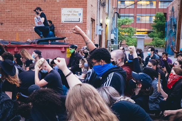 A group of people are gathered in an outdoor alleyway, raising their fists in a sign of protest. Many are wearing masks, and some wear bandanas or hats. The background shows a brick building with a sign and some greenery.