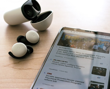 Wireless earbuds with a charging case are placed on a wooden surface next to a smartphone displaying news articles. The earbuds are black and white, and the phone shows text and images related to news headlines.