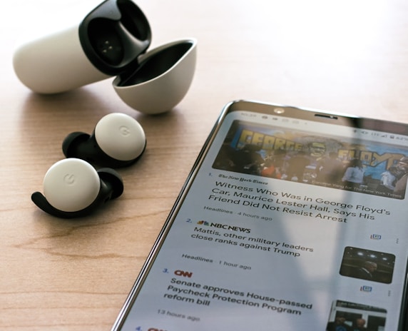 Wireless earbuds with a charging case are placed on a wooden surface next to a smartphone displaying news articles. The earbuds are black and white, and the phone shows text and images related to news headlines.