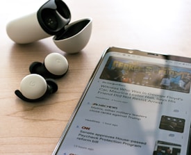 Wireless earbuds with a charging case are placed on a wooden surface next to a smartphone displaying news articles. The earbuds are black and white, and the phone shows text and images related to news headlines.