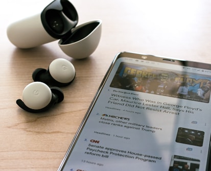 Wireless earbuds with a charging case are placed on a wooden surface next to a smartphone displaying news articles. The earbuds are black and white, and the phone shows text and images related to news headlines.