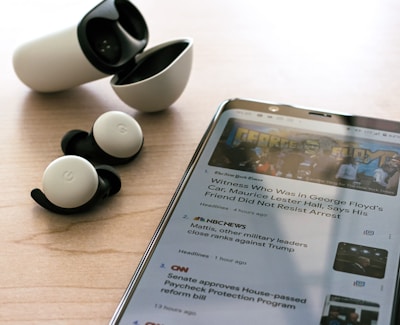 Wireless earbuds with a charging case are placed on a wooden surface next to a smartphone displaying news articles. The earbuds are black and white, and the phone shows text and images related to news headlines.