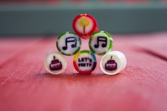 A red surface serves as the background for small, colorful candies arranged in a pyramid. Each candy has a unique design, including musical notes, a party hat, and the words 'LET'S PARTY' in bold letters.