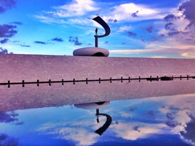 A striking sculpture on a large, minimalist white platform with a clear blue sky and dramatic clouds in the background. The foreground features a reflective body of water mirroring the structure and sky, creating a sense of symmetry and tranquility.