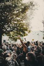 A group of people are gathered outside, many wearing masks, with their fists raised in the air as part of a demonstration or protest. The scene is set beneath a large, leafy tree with sunlight filtering through. Participants appear engaged and determined, holding various signs, and are surrounded by an urban environment.
