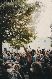 A group of people are gathered outside, many wearing masks, with their fists raised in the air as part of a demonstration or protest. The scene is set beneath a large, leafy tree with sunlight filtering through. Participants appear engaged and determined, holding various signs, and are surrounded by an urban environment.