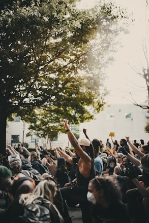 A group of people are gathered outside, many wearing masks, with their fists raised in the air as part of a demonstration or protest. The scene is set beneath a large, leafy tree with sunlight filtering through. Participants appear engaged and determined, holding various signs, and are surrounded by an urban environment.