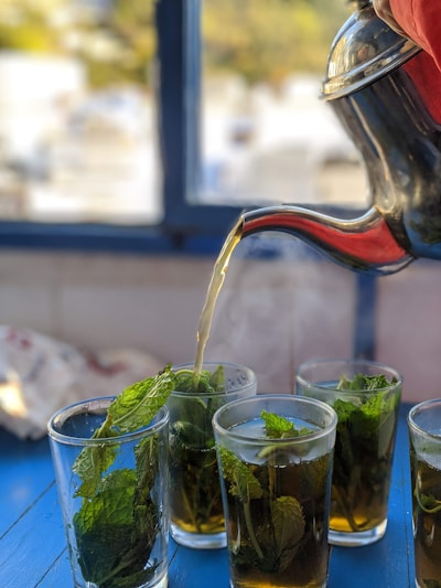 A stream of tea is being poured from a silver teapot into a clear glass filled with mint leaves, creating a refreshing beverage. The background features a window with soft-focus greenery outside. The setting appears to be in a bright, relaxed environment.
