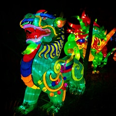 Vibrant, illuminated sculptures of mythical creatures with intricate patterns and bright colors. These figures are prominently set against a dark backdrop, likely in a nighttime setting, showcasing a dynamic mix of swirling lines and ornate details.
