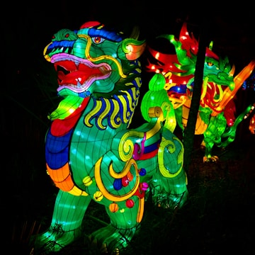 Vibrant, illuminated sculptures of mythical creatures with intricate patterns and bright colors. These figures are prominently set against a dark backdrop, likely in a nighttime setting, showcasing a dynamic mix of swirling lines and ornate details.