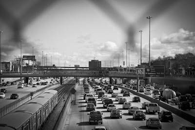 A crowded highway with heavy traffic is visible, with a parallel railway track next to it. Vehicles fill multiple lanes, and a train is visible on the tracks. Overhead, a bridge crosses the highway. Street signs and lamp posts are visible, and there are some trees and buildings in the background under a cloudy sky.