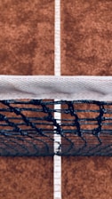 Close-up of a tennis racquet string bed highlighting the flat strings under natural sunlight on a clay court.