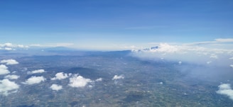 An aerial view of a sprawling landscape with scattered clouds casting shadows on the terrain below. A mountain range can be seen in the distance beneath a clear blue sky with minimal cloud coverage. The terrain appears mostly flat with patches of greenery and some urban development visible.
