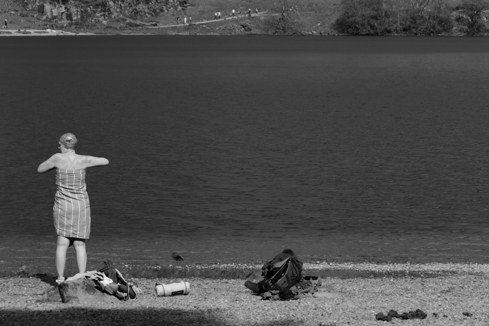 grayscale photo of person sitting on rock near body of water