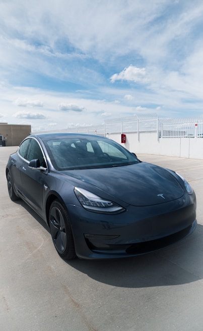 A sleek, dark-colored Tesla car is parked on a concrete surface under a partly cloudy sky. The car is facing slightly to the left, and the surrounding area is an open space with some fencing and a small red utility box on a white wall in the background.