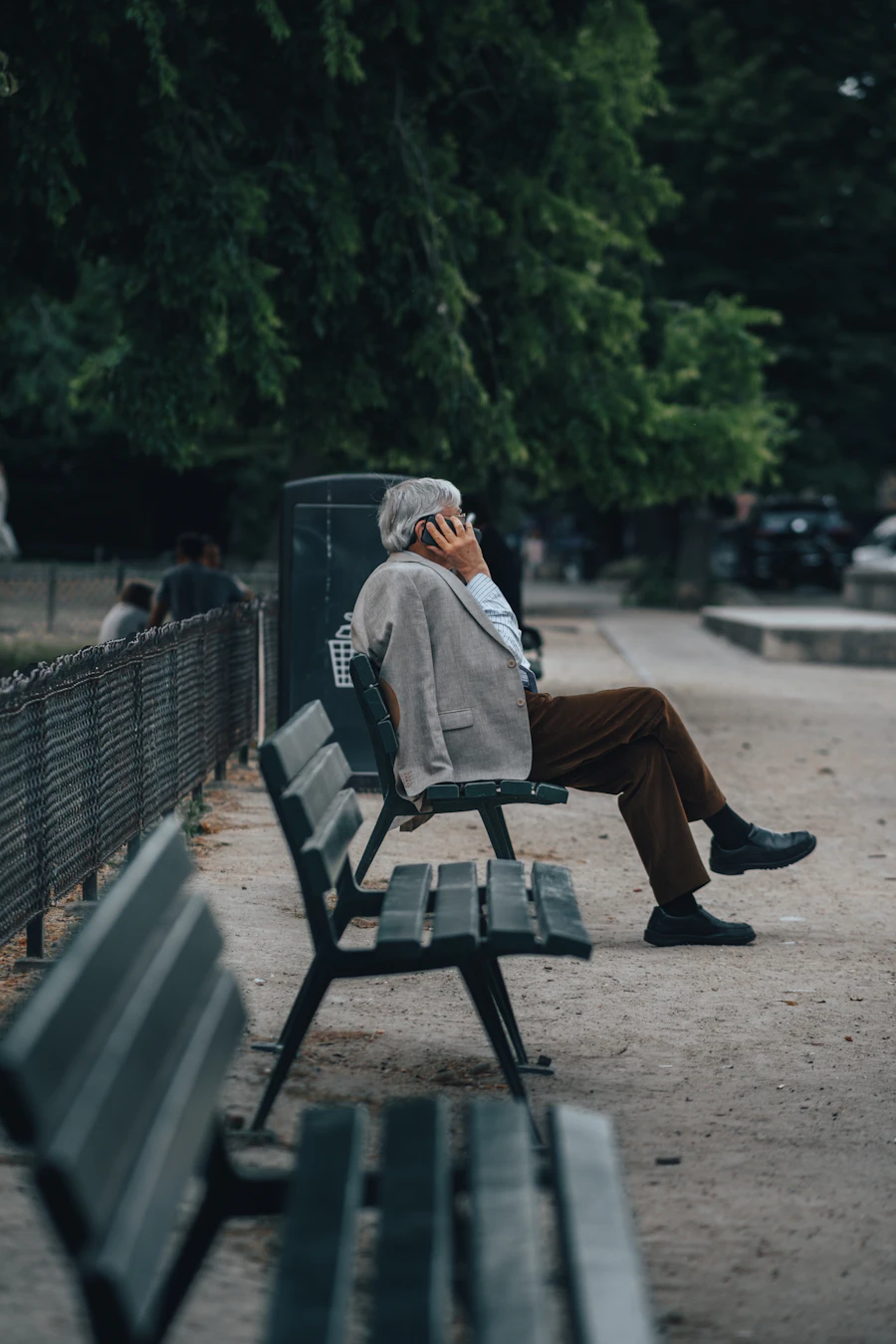 A man in a brown jacket sitting outside on a bench, holding a phone.