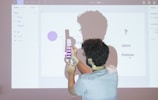 A person is standing in front of a projected screen, holding a color guide with several shades, while appearing to interact with the projected content. The screen displays various design elements, including text and color samples. The lighting creates a noticeable shadow of the person on the screen.