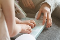 Two hands are gently touching a gift wrapped in light blue paper with decorative polar bears. The scene suggests a moment of sharing or gifting between two people, with one possibly guiding or helping the other. Part of an open book with pictures is visible in the background, suggesting an intimate, cozy setting.