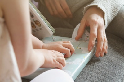 Two hands are gently touching a gift wrapped in light blue paper with decorative polar bears. The scene suggests a moment of sharing or gifting between two people, with one possibly guiding or helping the other. Part of an open book with pictures is visible in the background, suggesting an intimate, cozy setting.