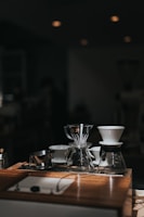 A collection of coffee brewing equipment is arranged neatly on a wooden counter. The setup includes glass coffee drippers, a metal measuring cup, and a glass carafe. The lighting creates a warm and inviting atmosphere, with soft shadows and highlights on the wooden surface.