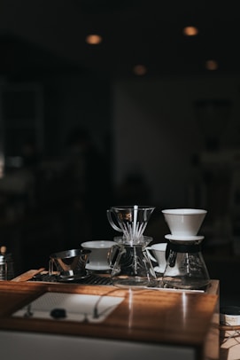 A collection of coffee brewing equipment is arranged neatly on a wooden counter. The setup includes glass coffee drippers, a metal measuring cup, and a glass carafe. The lighting creates a warm and inviting atmosphere, with soft shadows and highlights on the wooden surface.