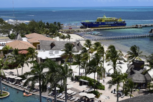 A vibrant coastal resort area with a large yellow and blue ferry docked at a pier, surrounded by clear blue waters. Palm trees line the edges of a swimming pool where people relax on lounge chairs, enjoying the sunny day. Thatched-roof buildings and a sandy shoreline contribute to a tropical ambiance.