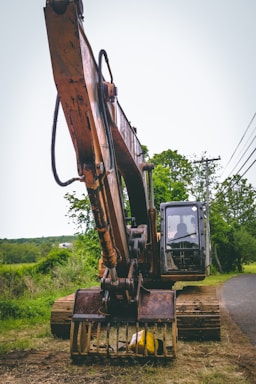 A large excavator with a rusty appearance stands on a grassy area near a road. Its arm and claw-like bucket are prominently displayed. The backdrop features lush greenery, trees, and a clear sky. A few power lines run parallel to the road.