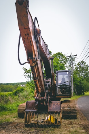 A large excavator with a rusty appearance stands on a grassy area near a road. Its arm and claw-like bucket are prominently displayed. The backdrop features lush greenery, trees, and a clear sky. A few power lines run parallel to the road.