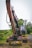 A large excavator with a rusty appearance stands on a grassy area near a road. Its arm and claw-like bucket are prominently displayed. The backdrop features lush greenery, trees, and a clear sky. A few power lines run parallel to the road.