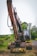 A large excavator with a rusty appearance stands on a grassy area near a road. Its arm and claw-like bucket are prominently displayed. The backdrop features lush greenery, trees, and a clear sky. A few power lines run parallel to the road.