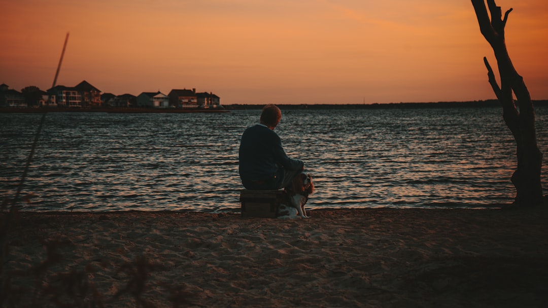 man in black jacket sitting on chair near body of water during sunset