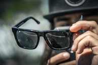 A person is holding a pair of stylish, dark-lensed sunglasses close to the camera. The person's fingers are visible, as well as a blurred background with greenery and indistinct shapes. The focus is primarily on the sunglasses, which reflect the light and surroundings subtly on their surface.