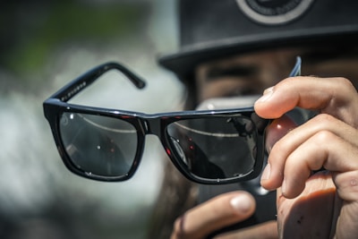 A person is holding a pair of stylish, dark-lensed sunglasses close to the camera. The person's fingers are visible, as well as a blurred background with greenery and indistinct shapes. The focus is primarily on the sunglasses, which reflect the light and surroundings subtly on their surface.