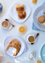 A selection of breakfast items including cups of coffee, croissants on white plates, lemon wedges, a few cookies on a white plate, and a small dish of cream. A blue-patterned bowl is partially visible, and the setting appears to be on a light-colored tabletop.