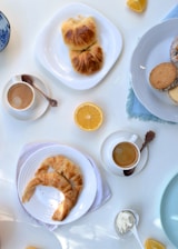 A selection of breakfast items including cups of coffee, croissants on white plates, lemon wedges, a few cookies on a white plate, and a small dish of cream. A blue-patterned bowl is partially visible, and the setting appears to be on a light-colored tabletop.