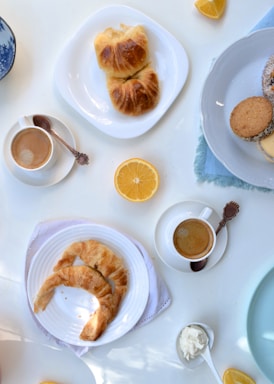 A selection of breakfast items including cups of coffee, croissants on white plates, lemon wedges, a few cookies on a white plate, and a small dish of cream. A blue-patterned bowl is partially visible, and the setting appears to be on a light-colored tabletop.