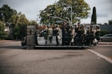 A group of SWAT officers is standing on a specialized armored vehicle at an intersection. The officers are dressed in tactical gear including helmets and protective vests. Trees and parked cars line the street, and a street sign indicates the location is Fountain Avenue.