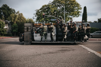 A group of SWAT officers is standing on a specialized armored vehicle at an intersection. The officers are dressed in tactical gear including helmets and protective vests. Trees and parked cars line the street, and a street sign indicates the location is Fountain Avenue.