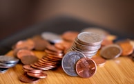 A collection of coins scattered on a wooden surface, with some coins stacked neatly in the center. The lighting emphasizes the metallic texture and colors of the coins, which include pennies, nickels, and dimes.