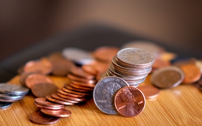 A collection of coins scattered on a wooden surface, with some coins stacked neatly in the center. The lighting emphasizes the metallic texture and colors of the coins, which include pennies, nickels, and dimes.