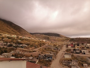 A rural community nestled in a mountainous area under a cloudy sky, featuring dirt roads and various small houses scattered across the landscape. The terrain is hilly and sparsely vegetated with patches of greenery.