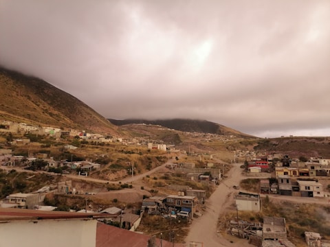 A rural community nestled in a mountainous area under a cloudy sky, featuring dirt roads and various small houses scattered across the landscape. The terrain is hilly and sparsely vegetated with patches of greenery.